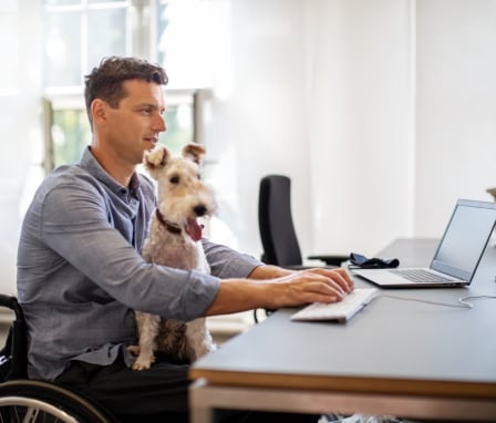 Man in wheelchair typing on computer with dog in lap